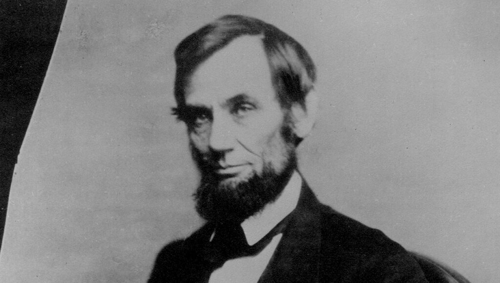 Abraham Lincoln, wearing his signature beard and dressed in a suit, is seated for a formal portrait. He looks off to his left. He is photographed from the legs up.
