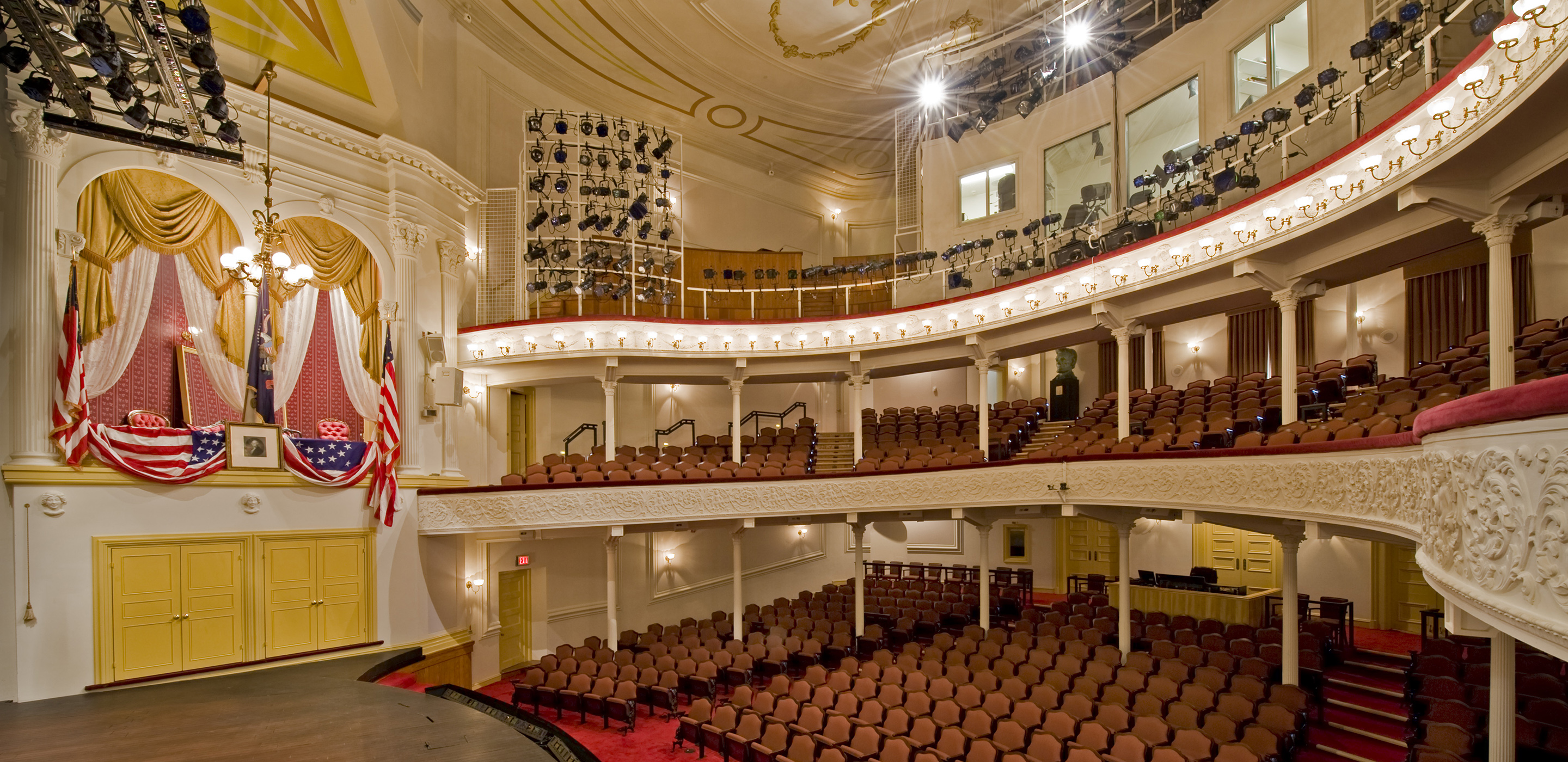 A side view of the stage and seating at Ford’s Theatre. On the left is the Presidential Box with an American flag, a framed picture of George Washington and American flag bunting draped over the box.