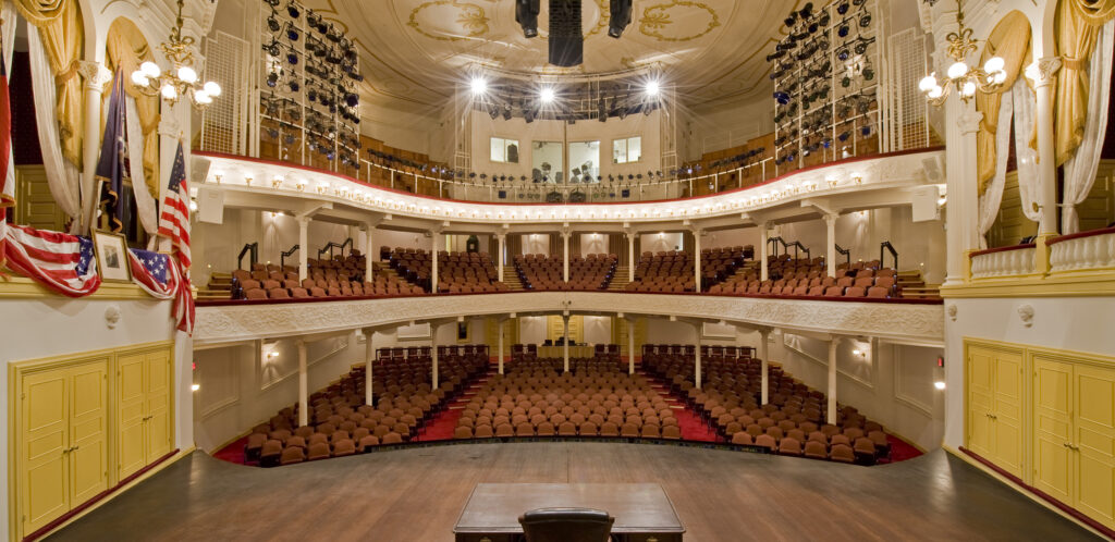 The view from the Ford’s Theatre stage looking out to the audience. To the left of the stage is the President Box with an American flag, a framed picture of George Washington and American flag bunting draped over the box. To the right is another box with yellow and white curtains. In the center of the stage is a wooden desk. The view includes two levels of seating and rows of lighting equipment on the third level.