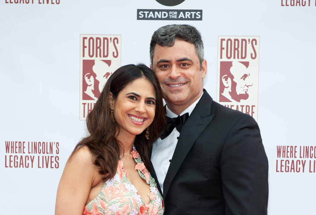 A man and a woman smile for the camera. Behind them is a backdrop covered in Ford's Theatre logos.