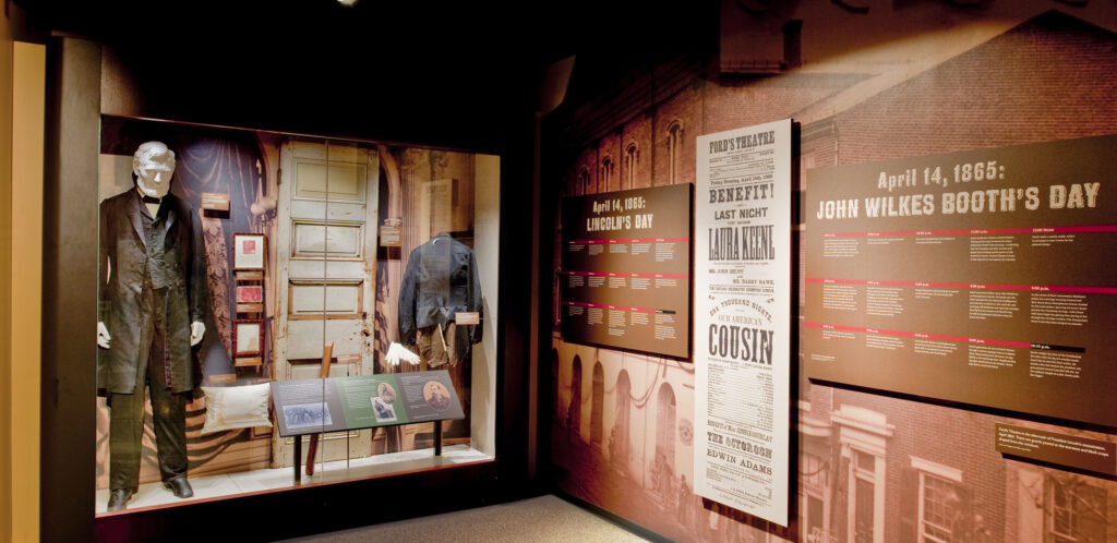 A display case with the black suit Lincoln was wearing the night of his assassination, the door leading to the theatre box and other relics from Ford’s Theatre. On the wall to the right of the display case is a timeline of Lincoln’s day on the date of the assassination, a large reproduction of the playbill for “Our American Cousin” and a timeline of Booth’s day for the date of the assassination.