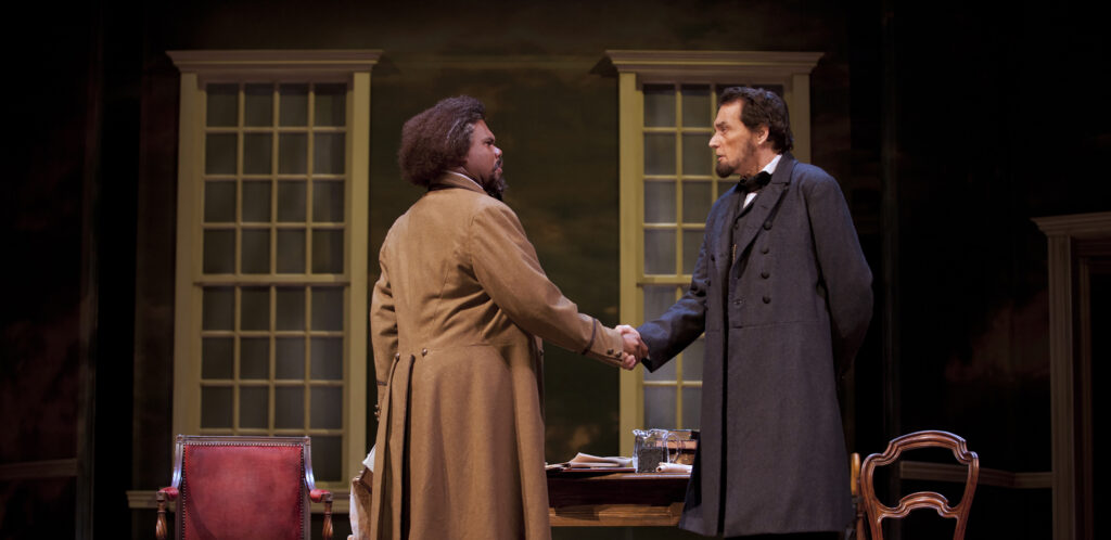 Actors portraying Frederick Douglass and Abraham Lincoln shake hands and talk while standing in front of a table and two closed windows.