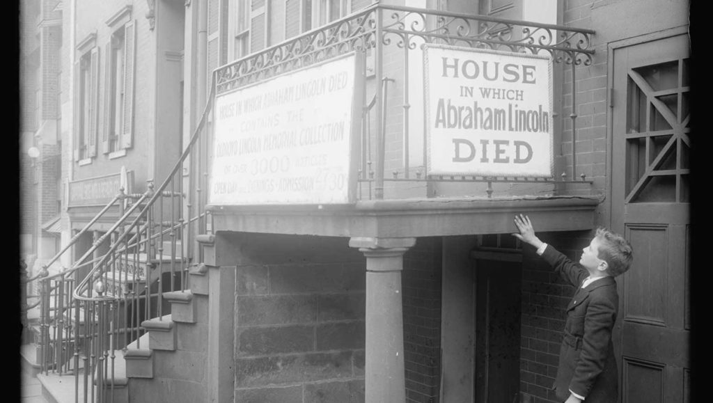 Black and white photograph of a young boy pointing to a sign in front of a house that reads "House in which Abraham Lincoln Died."