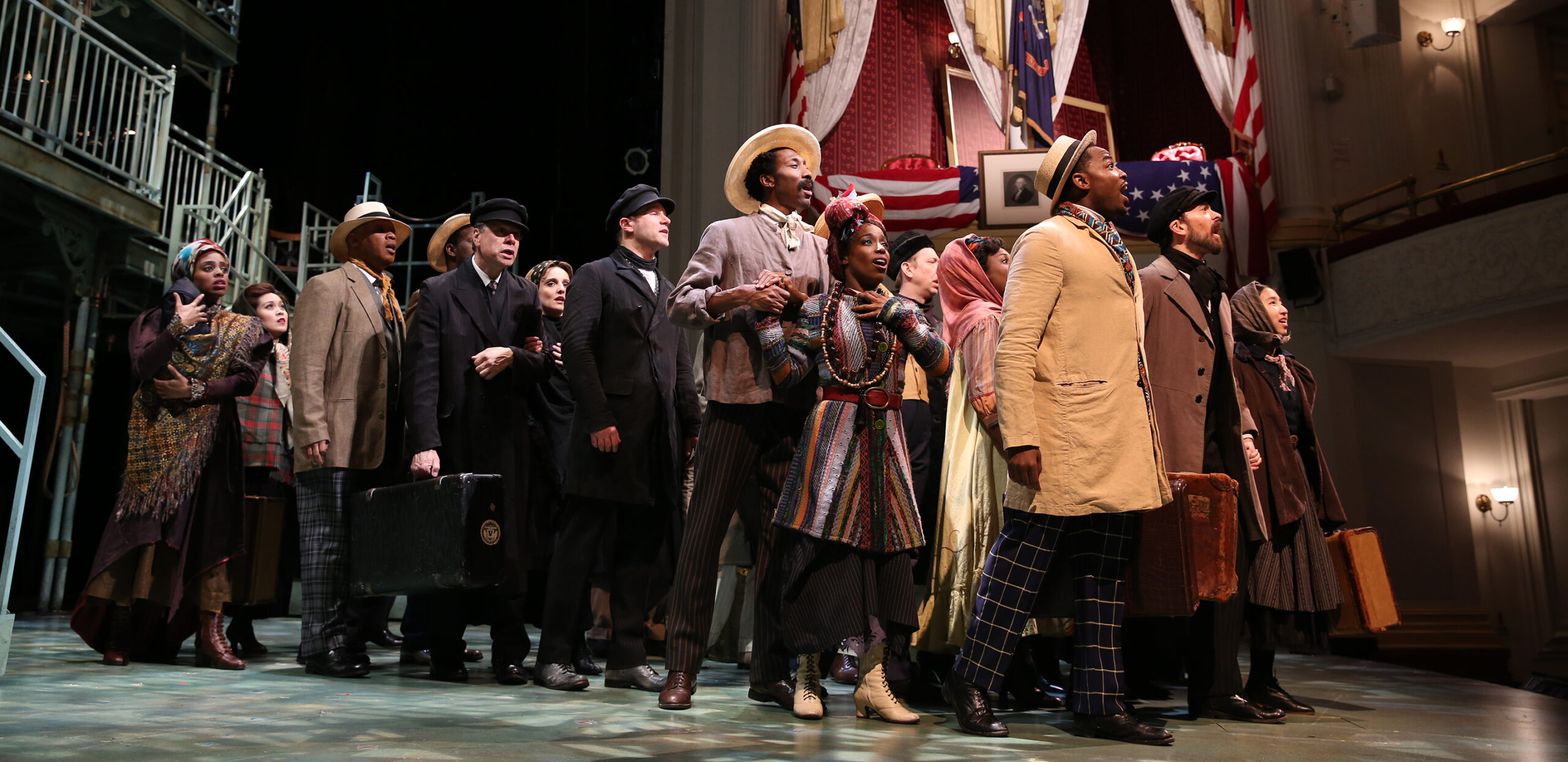 A large, multi-ethnic group of people dressed in early twentieth century clothes stand in a group and look to the right.