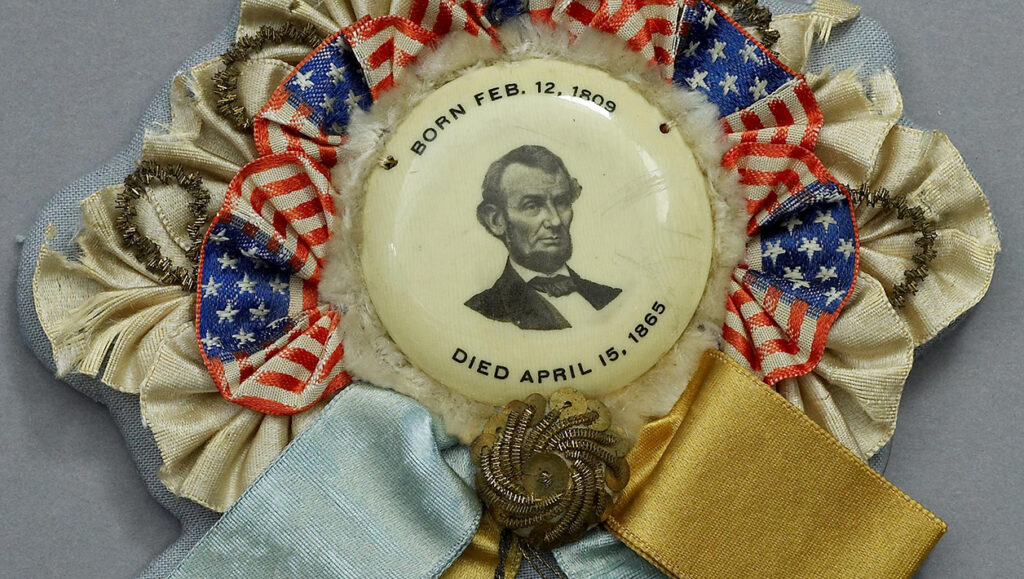 A mourning ribbon for President Lincoln with his portrait, birth and death dates in the center. Surrounding the portrait are small American flags. Hanging from the bottom are blue and hello ribbons and two decorative tassels.