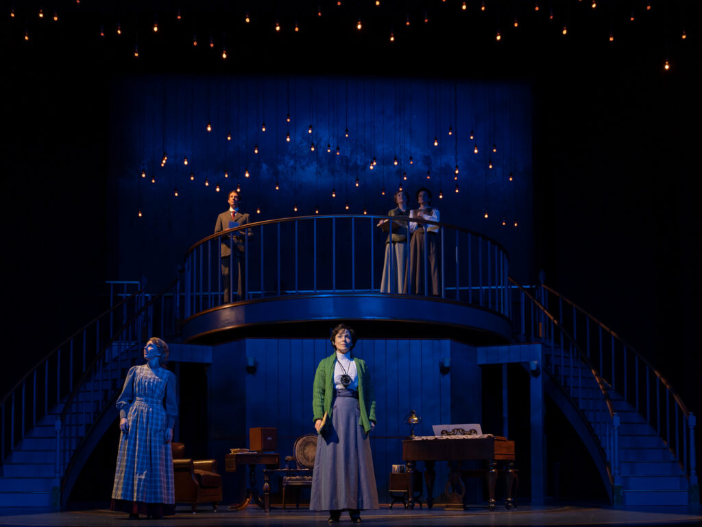 An actress stands center stage wearing a green sweater, 1910s white, long-sleeved blouse, a floor-length dark blue skirt and a period hearing aid. On the left, an actress wearing a fitted, floor-length apron in a checkered pattern gazes skyward. Above them three actors stand on a curved balcony platform also looking upward. Dozens of lightbulbs are suspended on black wires above the stage.