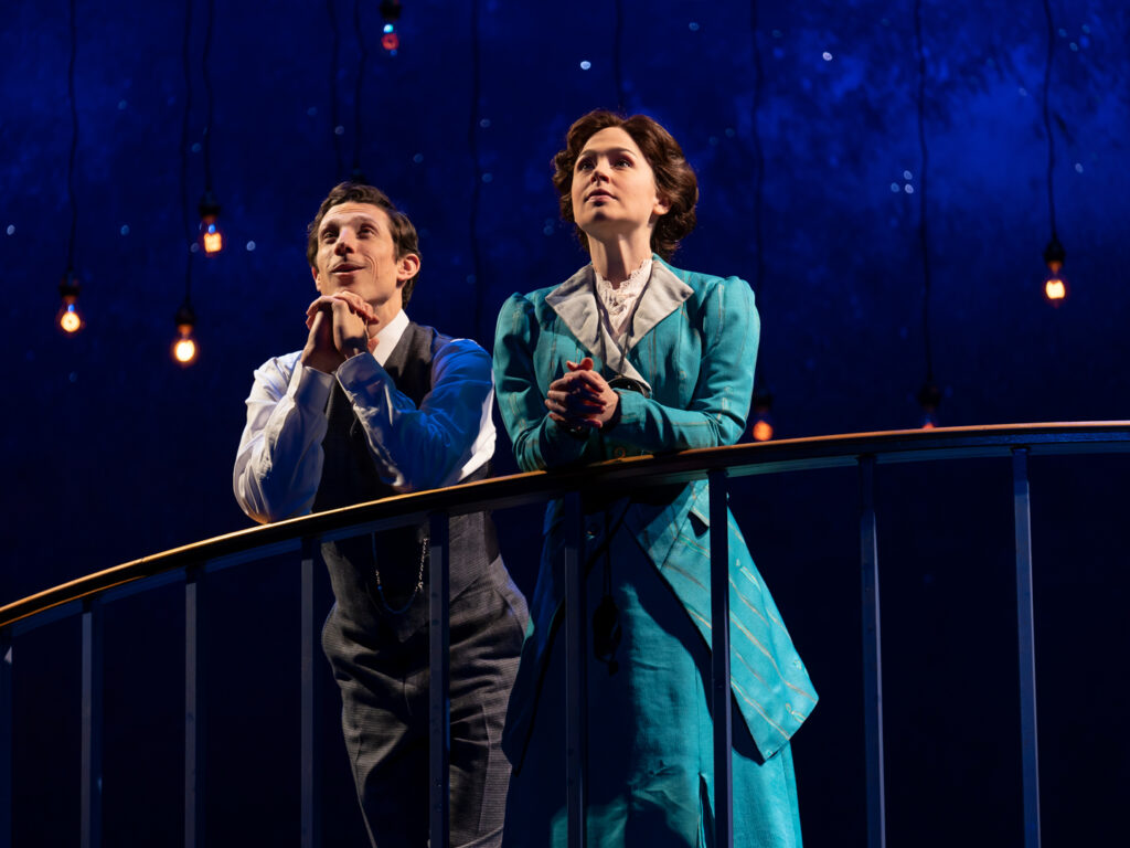 An actor wearing a 1910s white collared shirt, tweed vest and matching trousers stands with elbows resting on a balcony railing. On the right, an actress dressed in a teal jacket and matching floor-length dress, white collared shirt and a period hearing aid stands looking off into the distance. Above them, multiple lightbulbs are suspended on black wire.