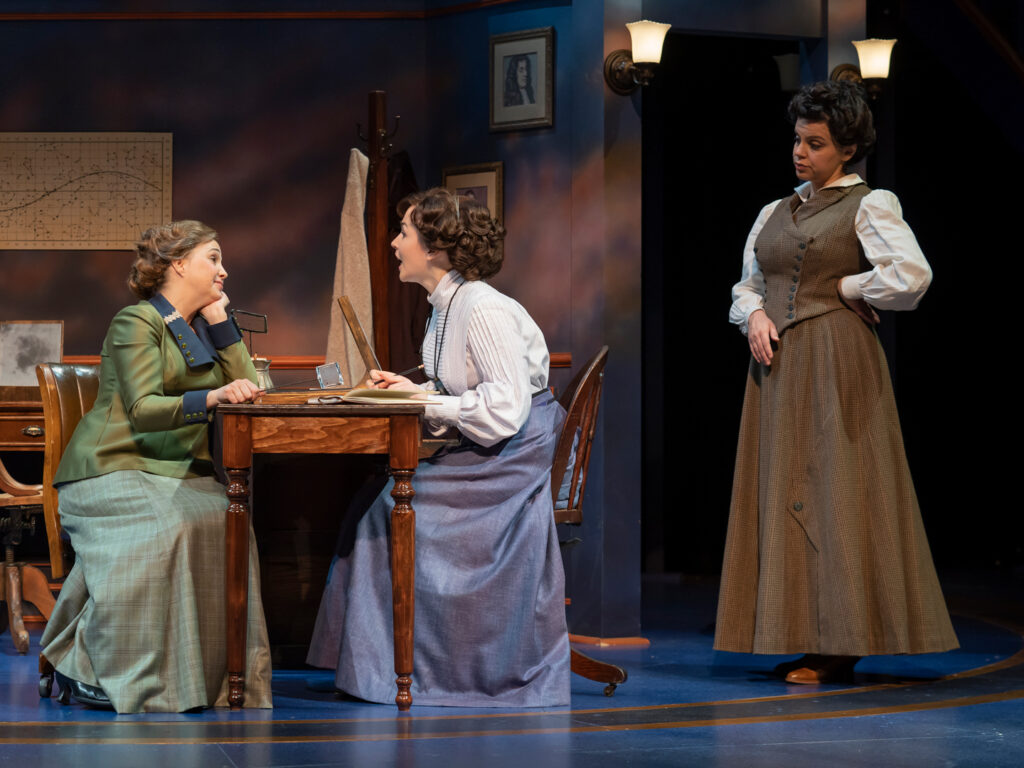 Two actresses wearing Victorian-era skirts and blouses sit on opposite sides of a turned-leg wooden desk. An actress portraying their supervisor stands behind them with one hand on her hip.