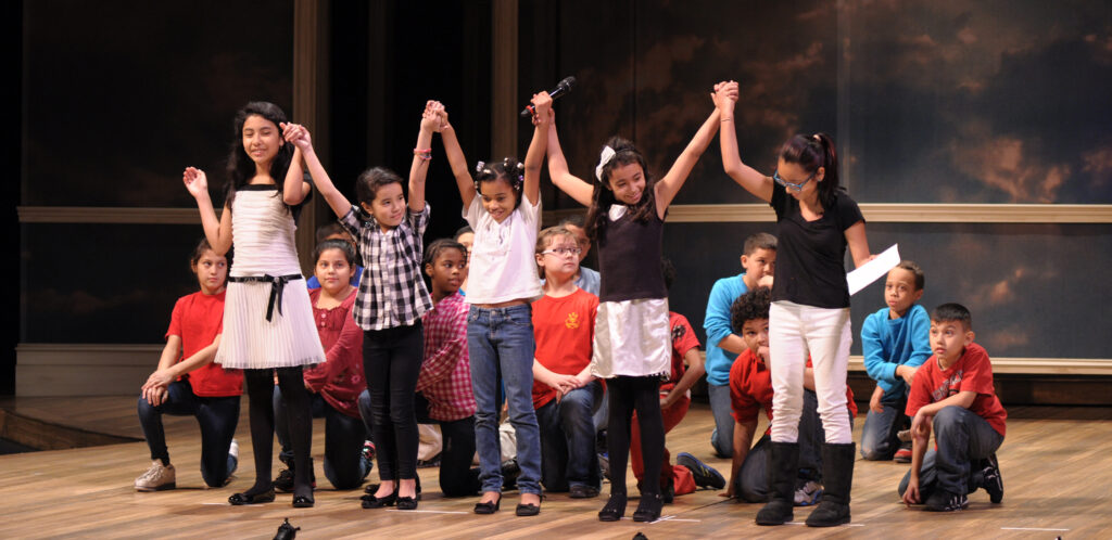 Students perform on stage as part of the Ford's theatre oratory festival. Five girls stand in front holding each other's hands above their heads. A dozen student kneel behind them.