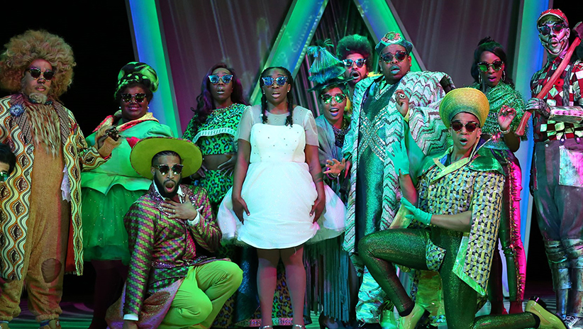 At center is Dorothy, wearing a white dress, silver shoes and mirrored sunglasses. She is surrounded by the residents of Emerald City, all dressed in shades of green and also wearing mirrored sunglasses. Everyone looks surprised.