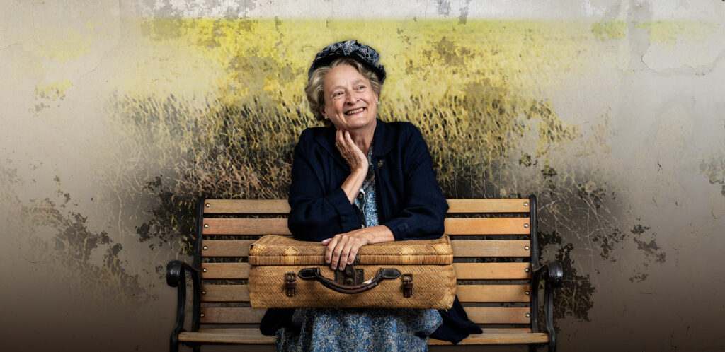 An elderly woman wearing a navy blue coat and matching hat, sits on a bench smiling with her suitcase on her lap.