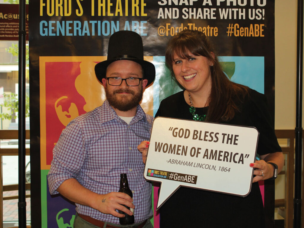 A man and woman stand next to each other and smile for the camera. The man wears a stovepipe hat and carries a beer bottle. The woman holds a cardboard sign that displays the words "God Bless the Women of America" Abraham Lincoln, 1864.