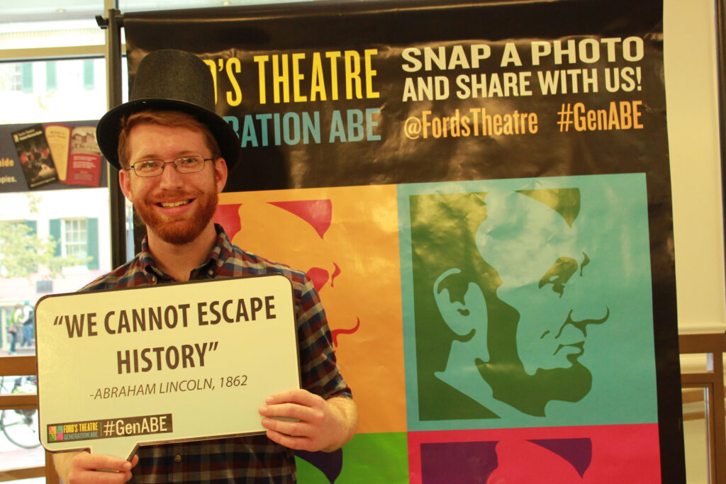 A man in a stovepipe hat smiles for the camera while holding a cardboard sign displaying the words "We cannot escape history" Abraham Lincoln, 1862.