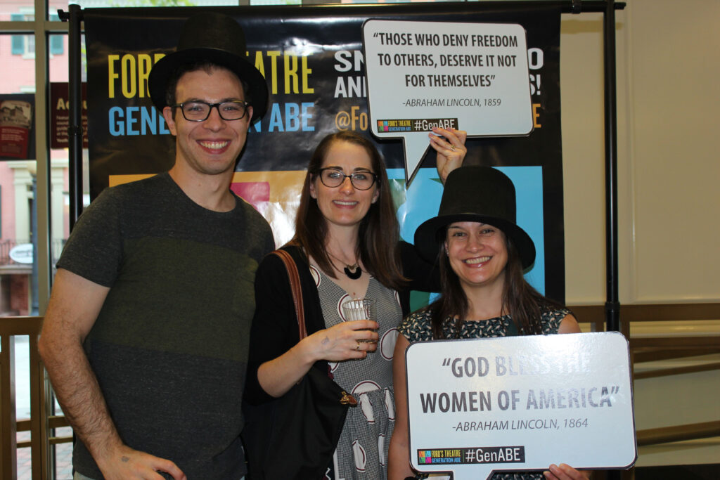 A man and two woman pose for a picture wearing stovepipe hats and holding cardboard signs displaying quotations from Abraham Lincoln.