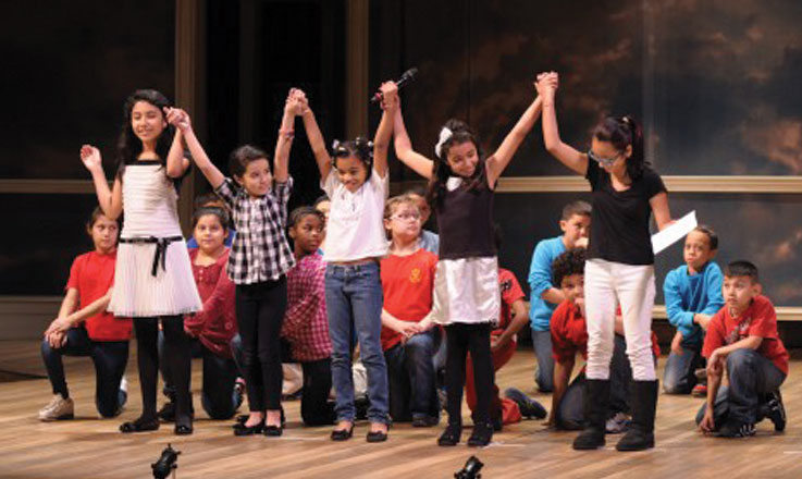 Students perform on stage as part of the Ford's theatre oratory festival. Five girls stand in front holding each other's hands above their heads. A dozen student kneel behind them.