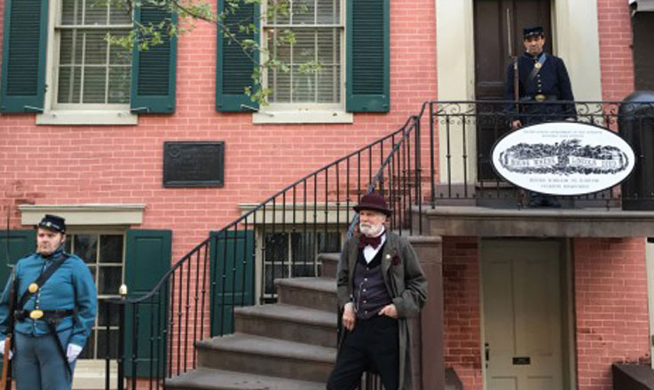 The chief of police stands in front of the stairs to the Petersen House, next to a union soldier. One more soldier stands guard at the top of the stairs, in front of the Petersen House front door.