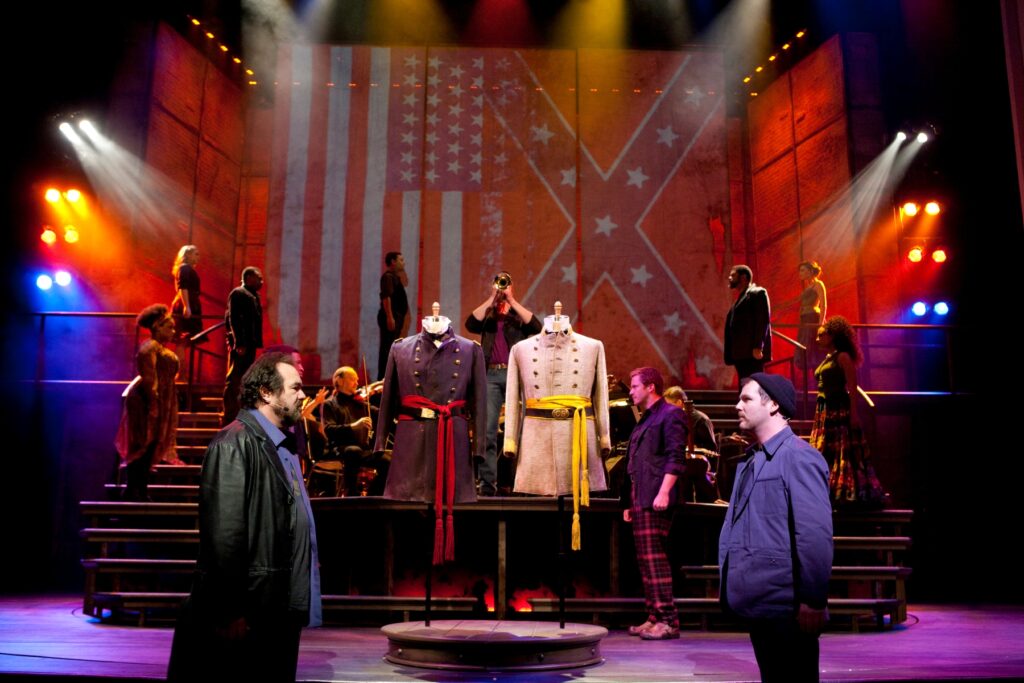 Four men face each other in pairs. Between them are a set of union and Confederate uniforms on dummies. At the rear of the stage a group of musicians plan, and the confederate and American flags are projected against the rear wall.