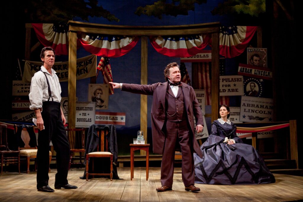 An actor portraying Stephen Douglas stands center stage and points his arm at an actor portraying Abraham Lincoln, who stands. An actress portraying the wife of Douglas sits off to the right side.