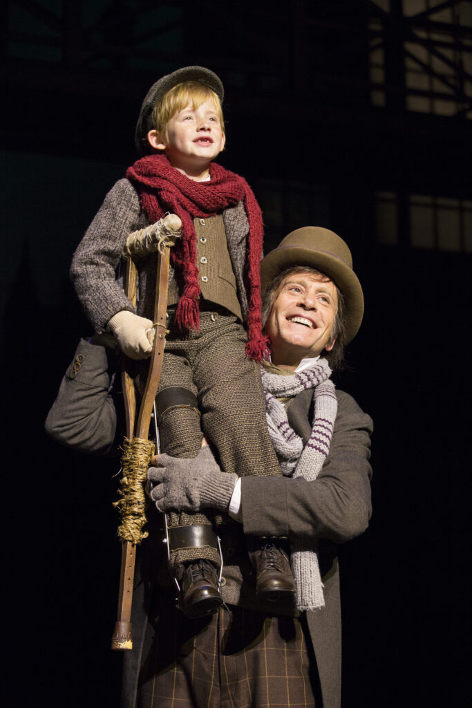 A man in Victorian era clothing holds up a small boy with a leg brace and crutch. Both are smiling.