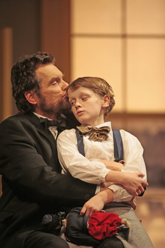 An actor portraying Abraham Lincoln holds an actor portraying his young son and kisses his forehead.