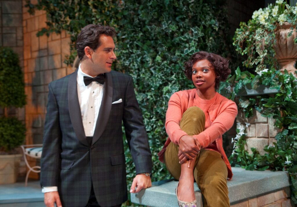 To the left stands a young white man in his thirties wearing a dark blue-and-green plaid suit with a black bowtie. He is leaning against a hip-height garden wall. Sitting on the wall is a young Black woman in a peach cardigan and brown pedal-pushers with her right knee tucked to her chest. The man is looking at her, while her focus is off to her left. The garden wall is covered in ivy and flowers behind them.
