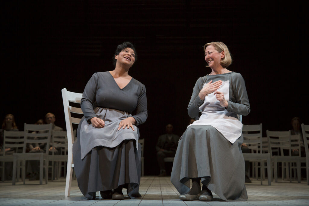 Two women dressed in gray sit in chairs on a stage and laugh together.
