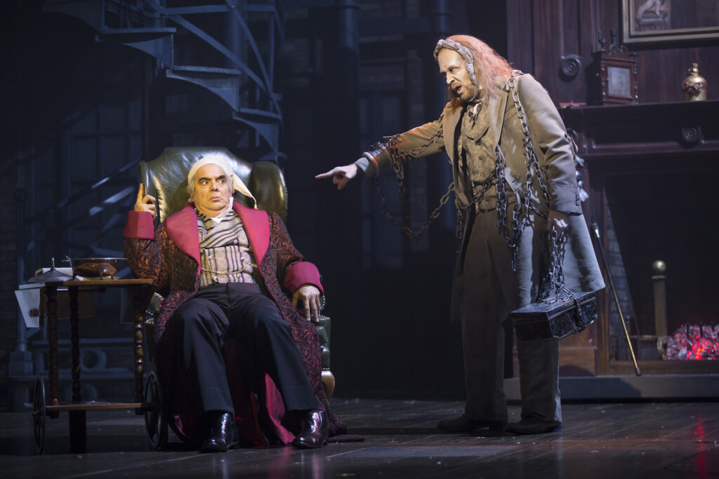 A ghostly man in ragged clothing and covered in chains points at a frightened man in his nightclothes who sits in a lounge chair.