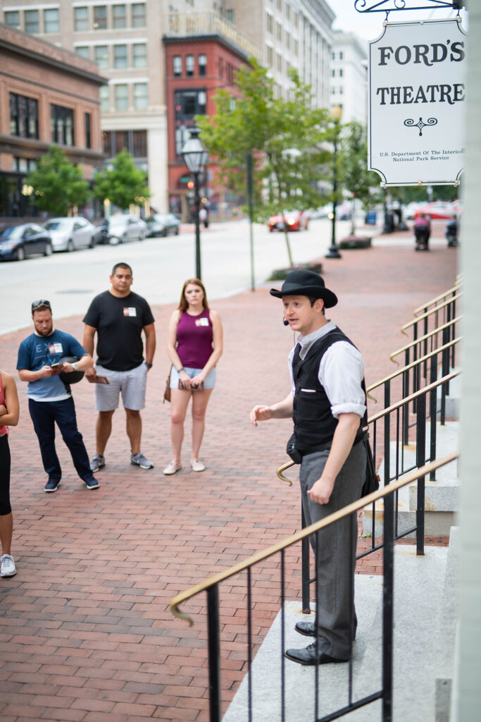 An actor dressed as Detective James McDevitt and wearing a microphone addresses a group of tour attendees on the steps of Ford’s Theatre.