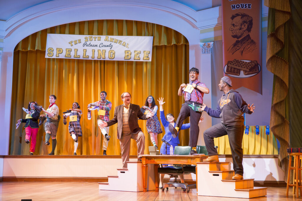 An actor dressed as a child in suspenders and a tie stands on a table and dances. Three adults dance around him. Behind them students dance on a raised stage. Above them is a sign that says "The 25th Annual Putman County Spelling Bee."