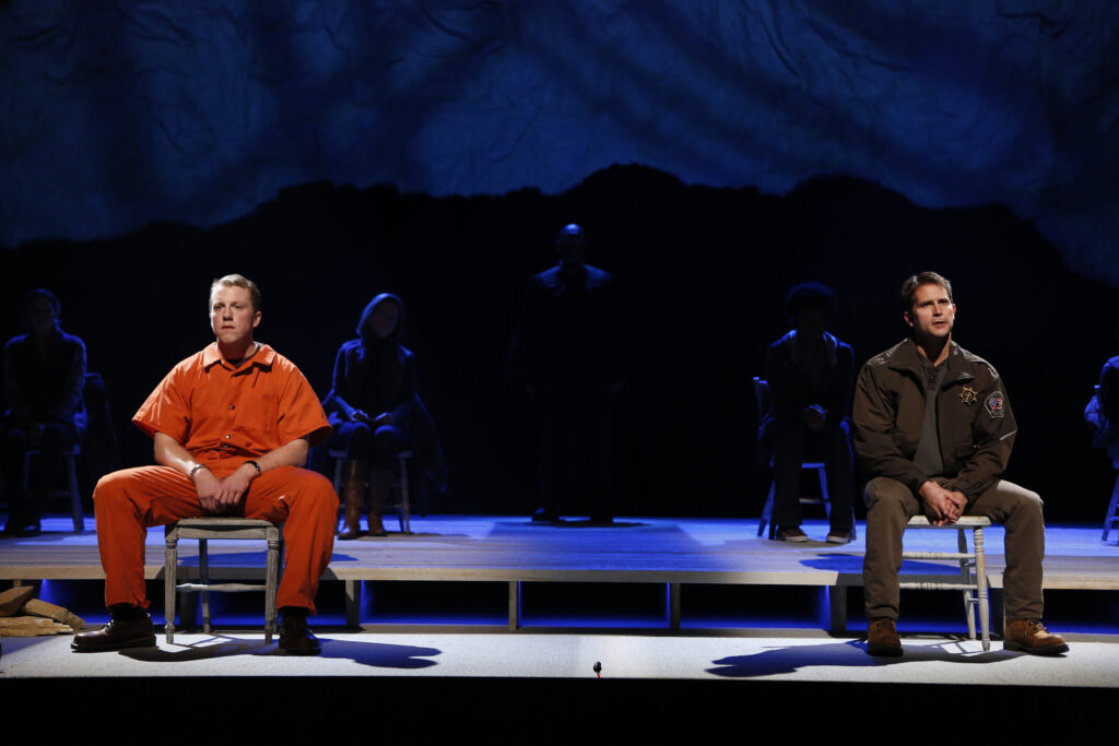 Two men, one dressed in a police uniform, the other in an orange prisoner's jumpsuit, sit far apart on a stage. Behind them other people sit or stand in shadow.