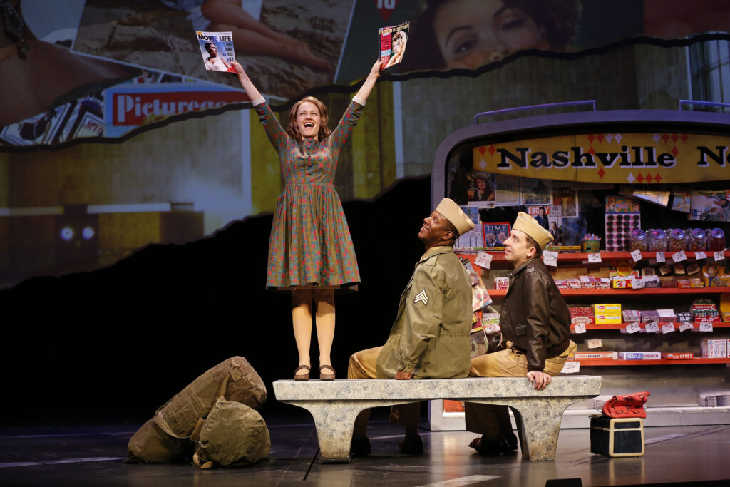 A young woman in a dress stands on a bunch and holds up a magazine in each hand. Two men in WWII era military uniforms sit on the bench and watch her. Behind them is a news stand with the words "Nashville News" written on a sign.
