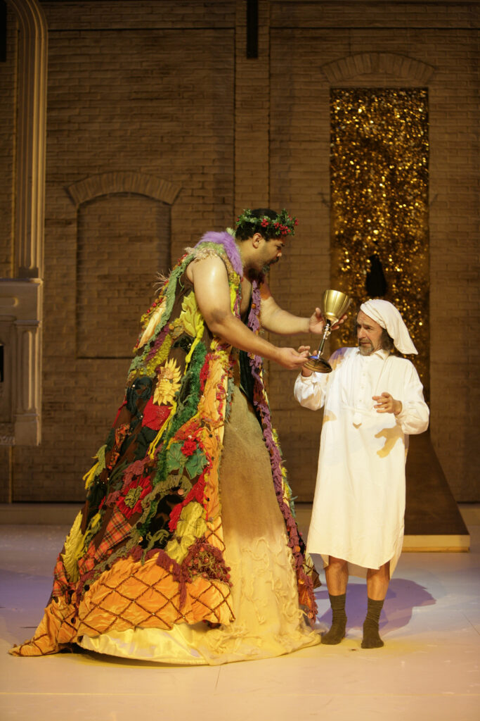 An overly tall man wearing a long patchwork gown and a holly wreath on his head hands a large goblet to a man in white nightclothes and a white sleeping cap.