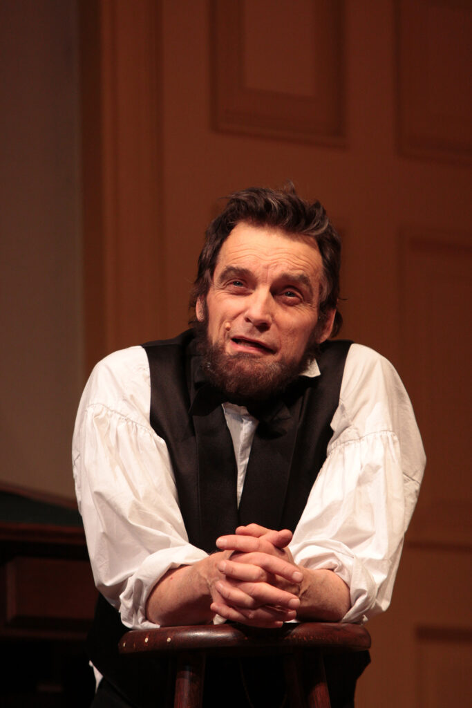 An actor portraying Abraham Lincoln leans forward onto a stool with his fingers intertwined and speaks.