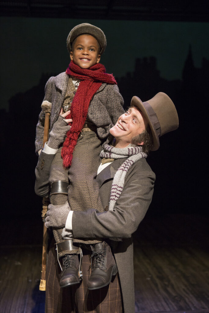 A man in Victorian clothing and a top hat holds a young boy with a crutch and leg brace up on his shoulder. They both smile.