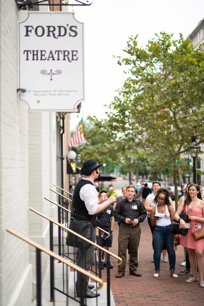 An actor dressed as Detective James McDevitt and wearing a microphone addresses a group of tour attendees on the steps of Ford’s Theatre.