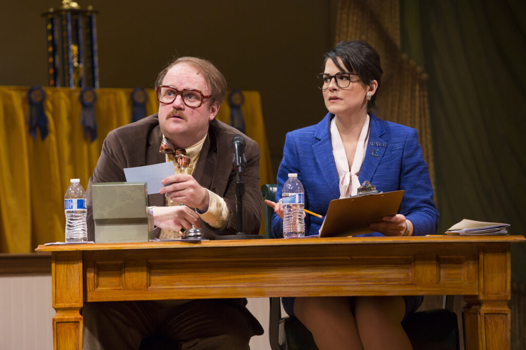 A man in a jacket and bowtie and a woman in a blue blazer sit at a table. The man holds a card in his hand, the woman holds a clipboard. Both look concernedly at something off stage.