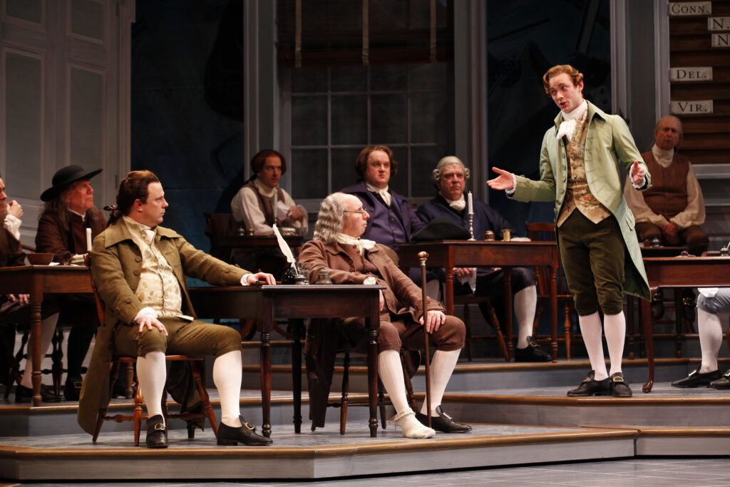 Actors portraying John Adams and Benjamin Franklin sit and listen to a man in a green coat and suit speaks. Behind them men sit at desks and watch.