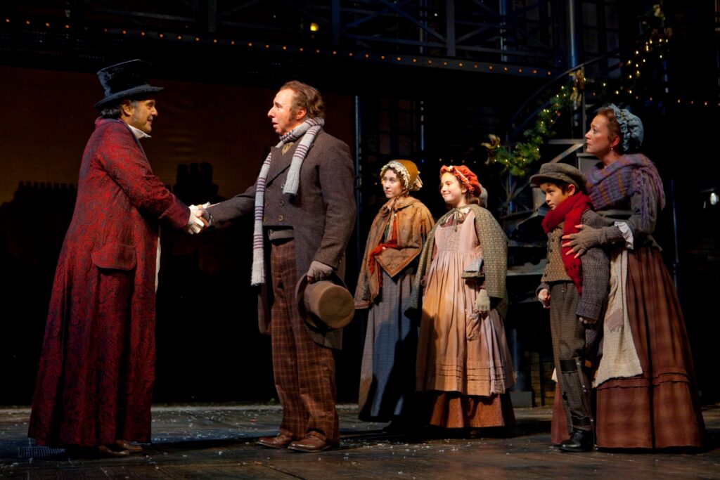 To the left a tall, older White man in a black top hat and a patterned red silk dressing-gown shakes hands with a middle-aged White man, who is wearing tweed pants, a gray goat, and a light blue scarf and holds a brown worn top hat in his left hand. The man in the red, Scrooge, is smiling while the other man, Bob Cratchit, looks perplexed. Behind Cratchit stand two young White girls in floor-length dresses and cloaks. Next to the girls is a middle-aged White woman in a floor-length skirt with an apron, gray fingerless gloves, and a plaid scarf with her hands on the shoulders of a young White boy who leans against her. The boy wears a tweed suit and cap and carries a cane in his left hand.