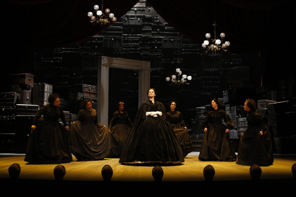 An actress portraying Mary Lincoln stands centerstage in a black gown and cape, and wearing white gloves. Around her in a semi-circle stand six curtseying women in black.