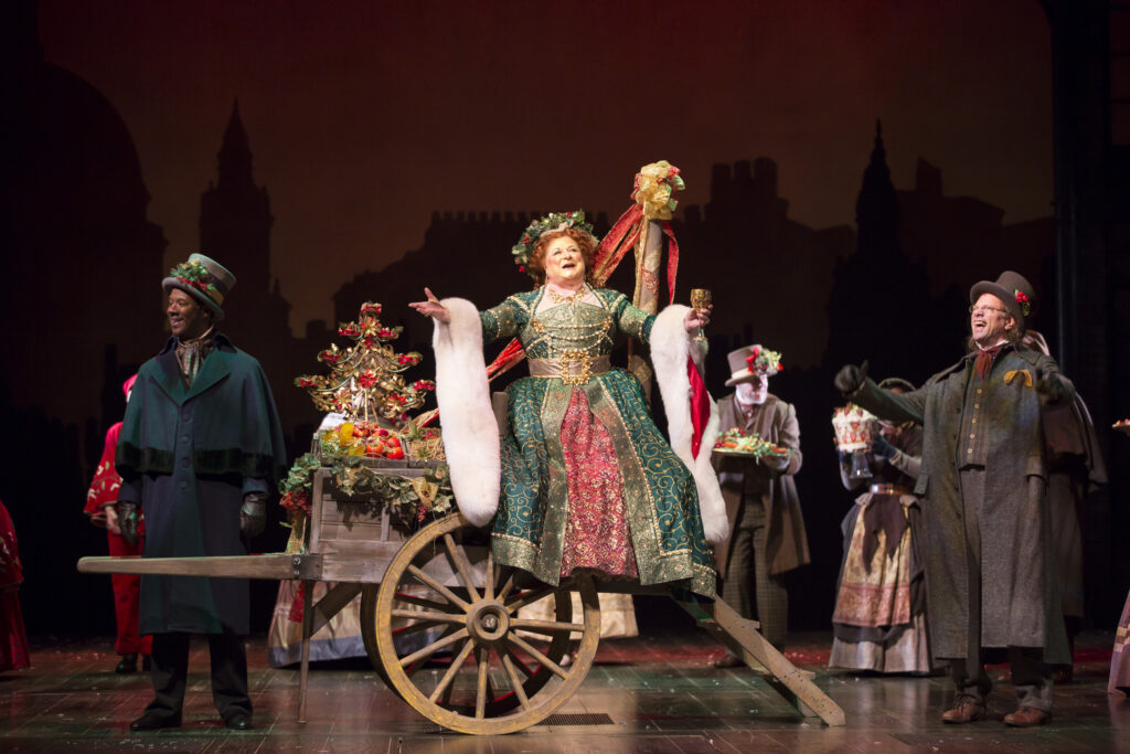 A woman in a green and red dress with a wreath on her head sits on a full wheelbarrow and holds up a goblet. Around her men and woman in holiday outfits sing.
