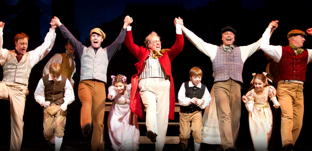 A group of five men dance and hold each other's hands up in the air while children run underneath.