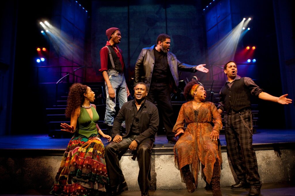 Six African-Americans sing on stage. Three stands and three sit.