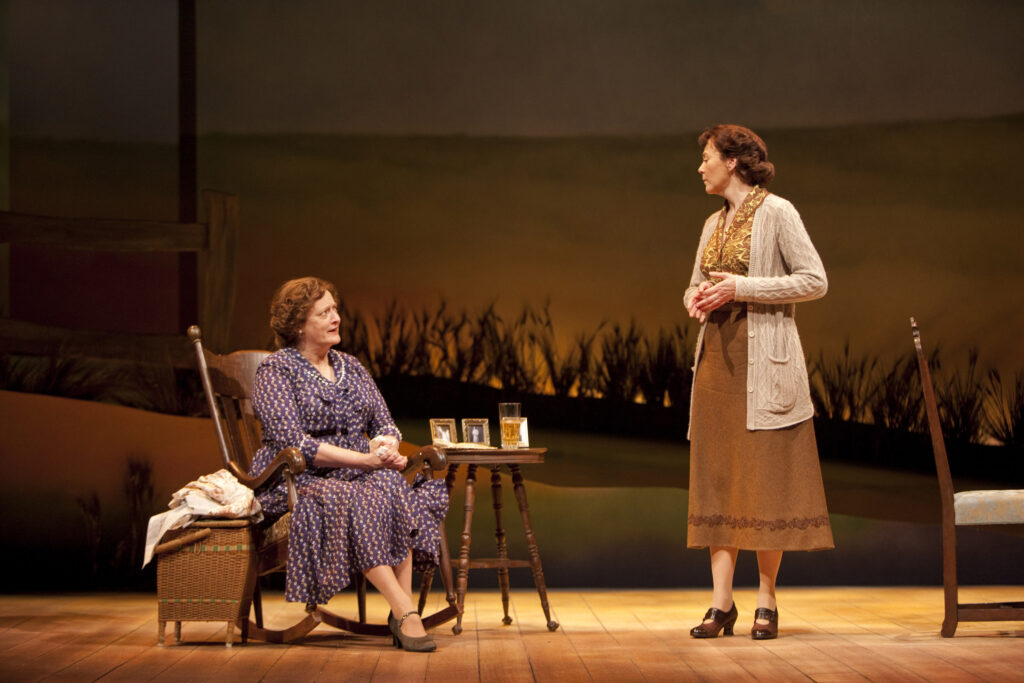 A woman in a purple dress sits on a rocking chair and speaks to a standing woman in a skirt, blouse and jacket.