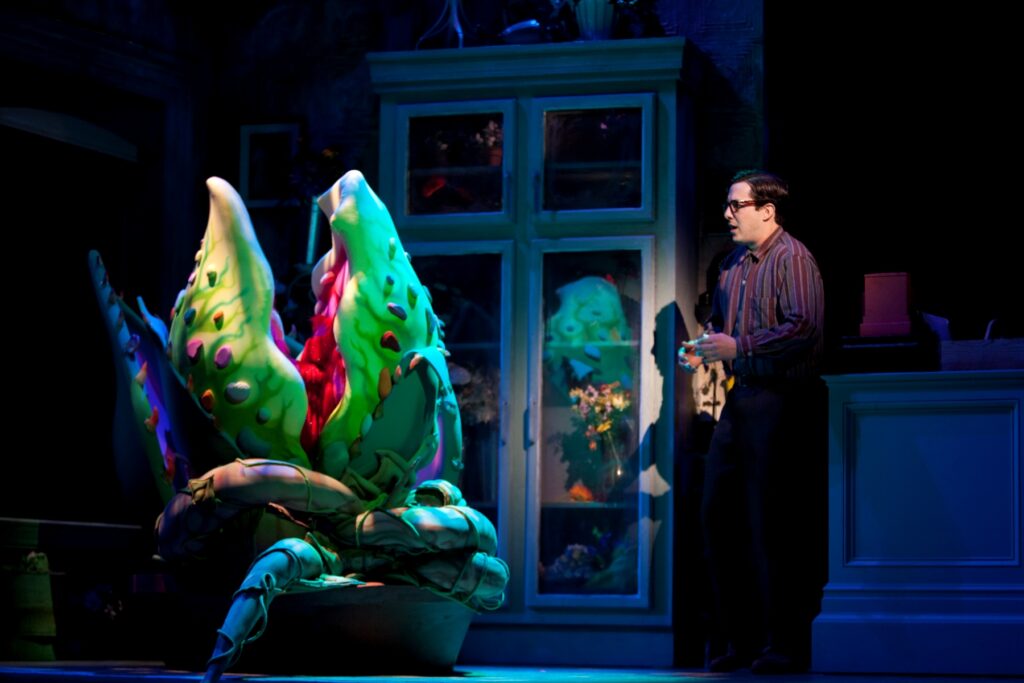 A man in glasses and a button down shirt stands in a bedroom and talks to a giant plant with a huge mouth in a pot.