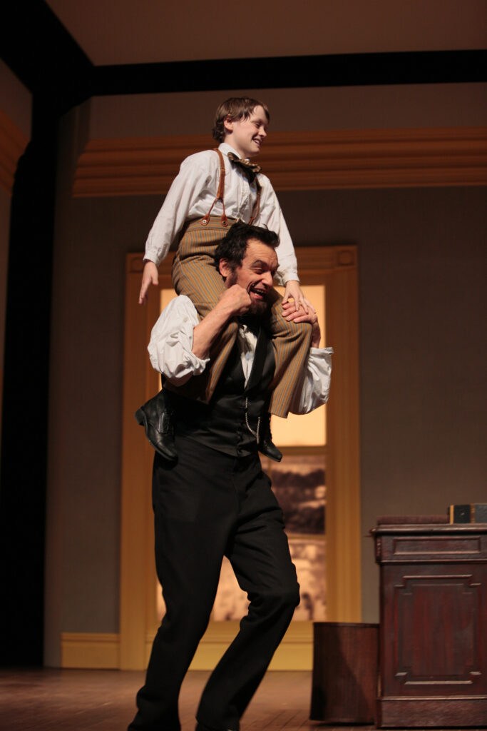 An actor portraying Tad Lincoln sits on the shoulders of an actor portraying Abraham Lincoln as he walks through an office. Both are smiling broadly.