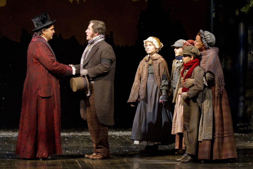 A man in Victorian nightclothes and top hat shakes the hand of a man in a long brown coat. Behind the man stand a group of children and an adult woman.