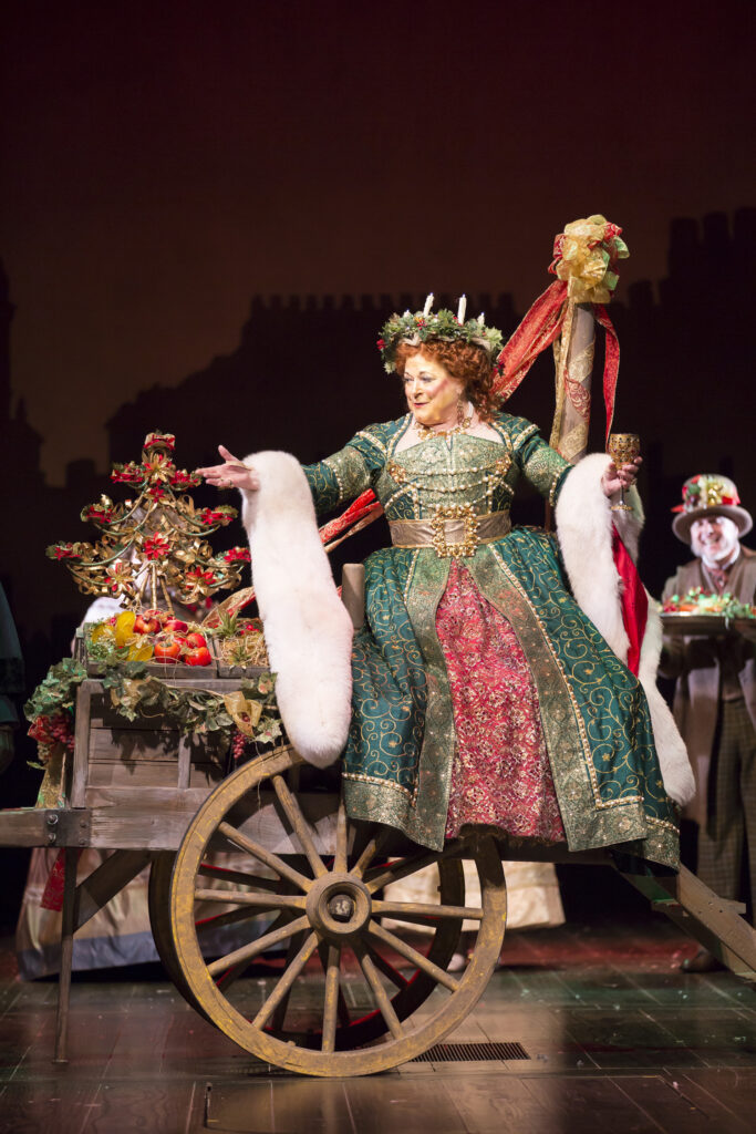 A woman in a green and red dress with a wreath on her head sits on a full wheelbarrow and holds up a goblet.