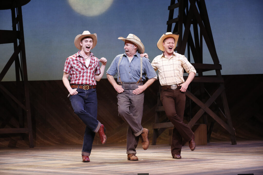 Three men wearing ten gallon hats all dance together in front of some oil derricks.