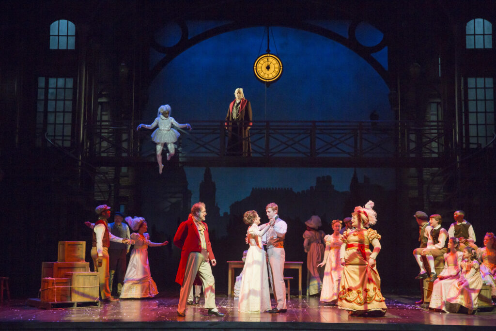 A group of men and women in fancy Victorian clothing dance on a stage. Above them, a woman in a white dress floats in the air, while a man in his nightclothes stands on a nearby catwalk and watches the dancers.