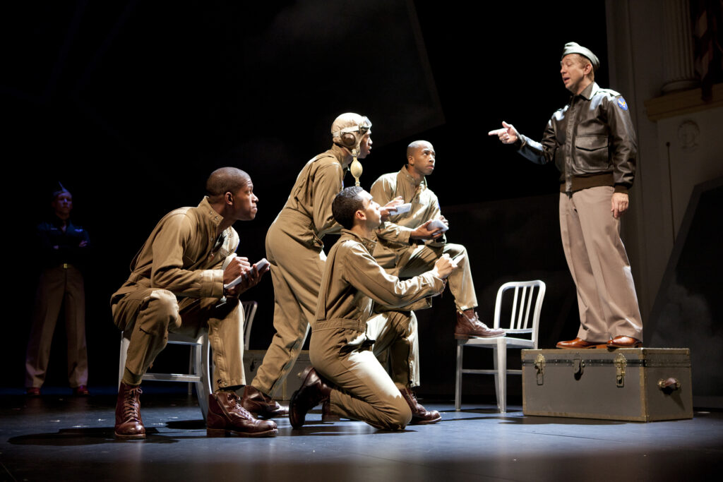 Four African-American men in khaki military uniforms squat or kneel in front of a white man in an aviator jacket standing on a trunk. They take notes while the man speaks to them.